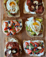 A variety of open-faced toasts topped with different combinations of ingredients, including fried eggs, avocado, sliced tomatoes, strawberries, blueberries, and almonds.