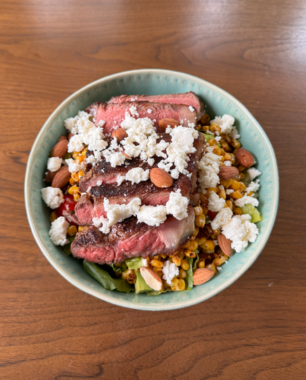 Close-up of a steak salad featuring sliced steak, Original Daily Crunch almonds, seasoned corn, and crumbled queso fresco, served in a green bowl.